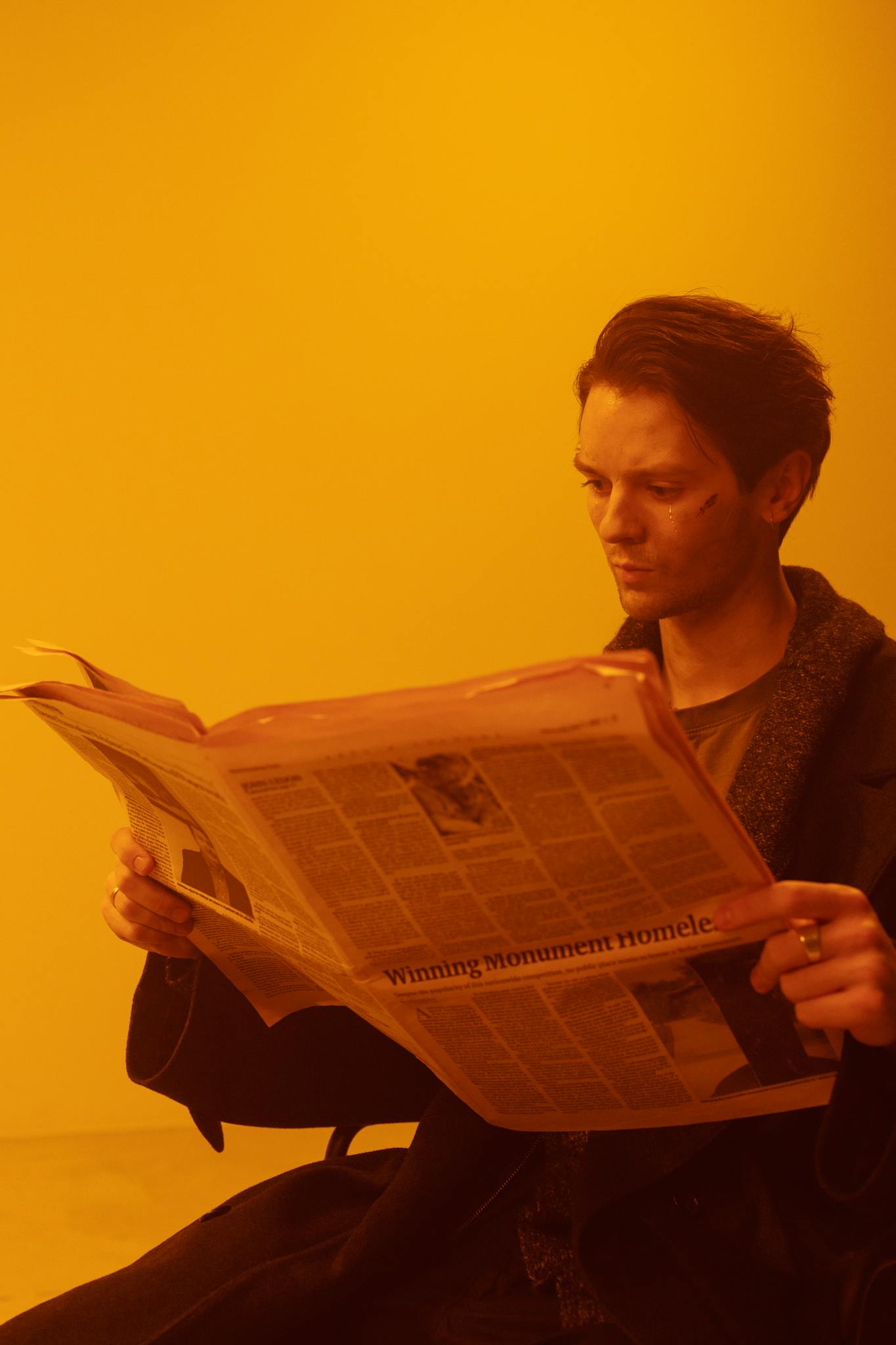 Adult man reading a newspaper in a warm, conceptual setting, wearing a coat.