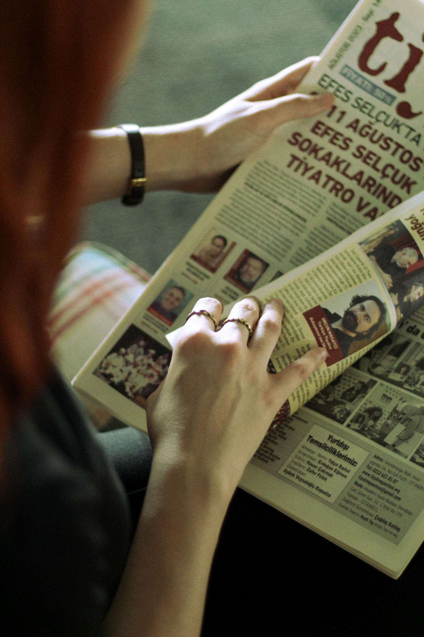 Close-up of a woman's hands reading a newspaper, seated indoors with selective focus.