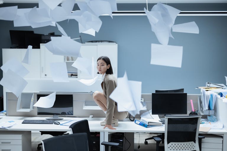 Woman perched on a desk in a chaotic office with papers flying, representing workplace stress.