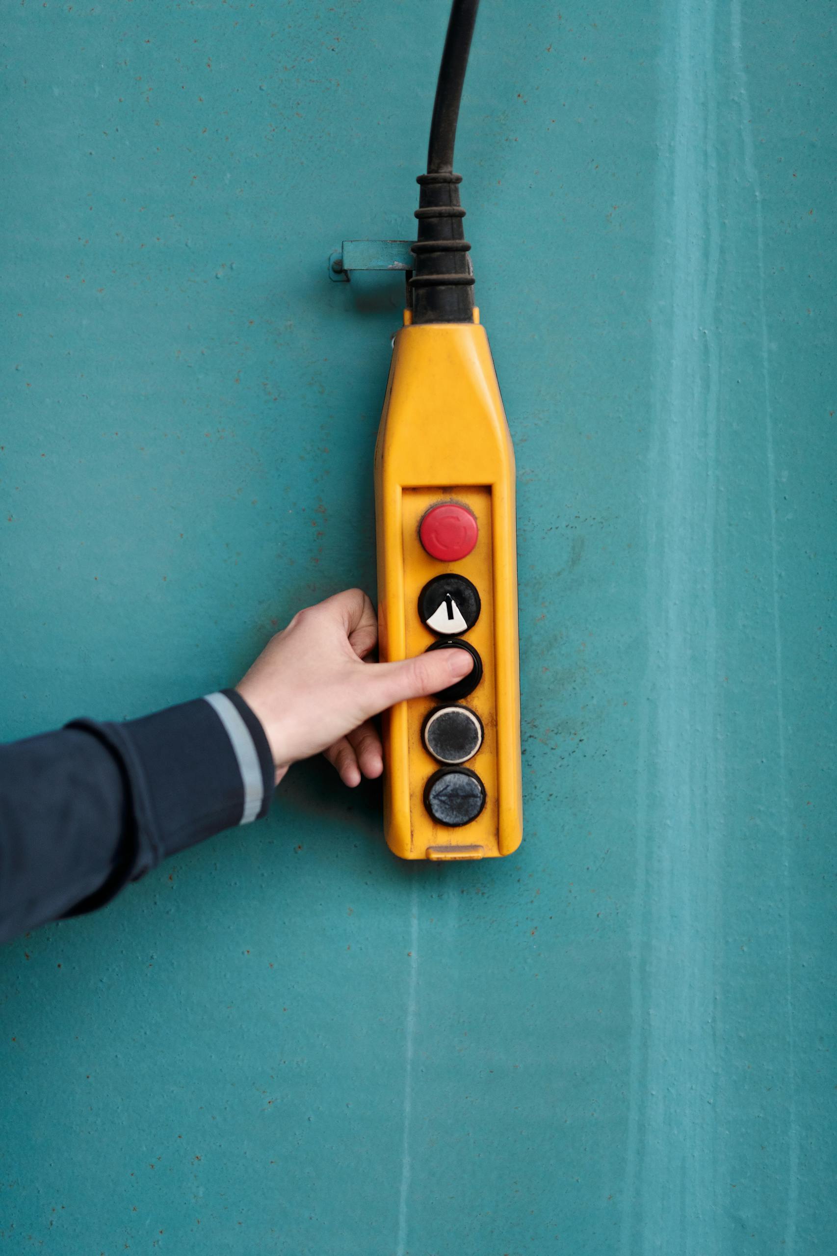 Close-up of a hand pressing a button on a yellow industrial control panel against a blue wall.