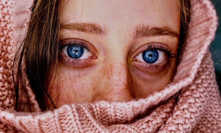 Intense close-up of a woman's blue eyes wrapped in a soft, pink knit scarf.
