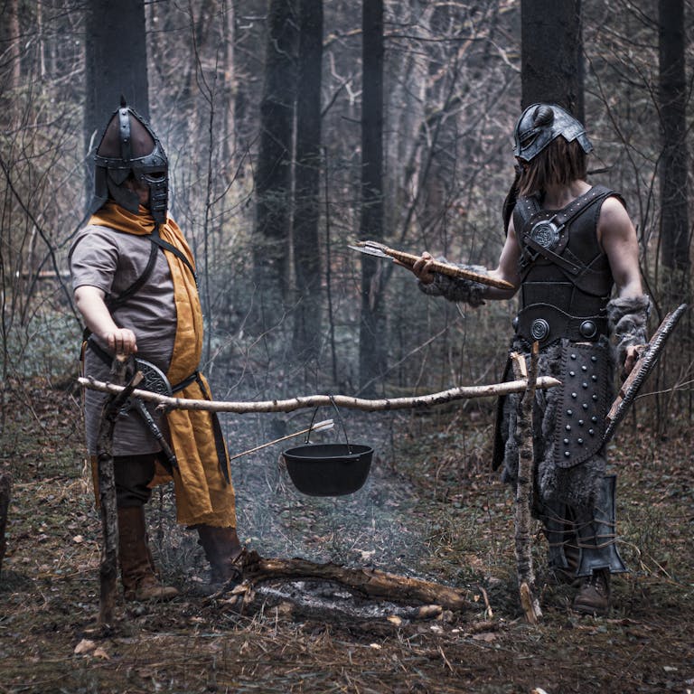 Two medieval reenactors cooking over a bonfire in a dense forest, dressed in historical armor.