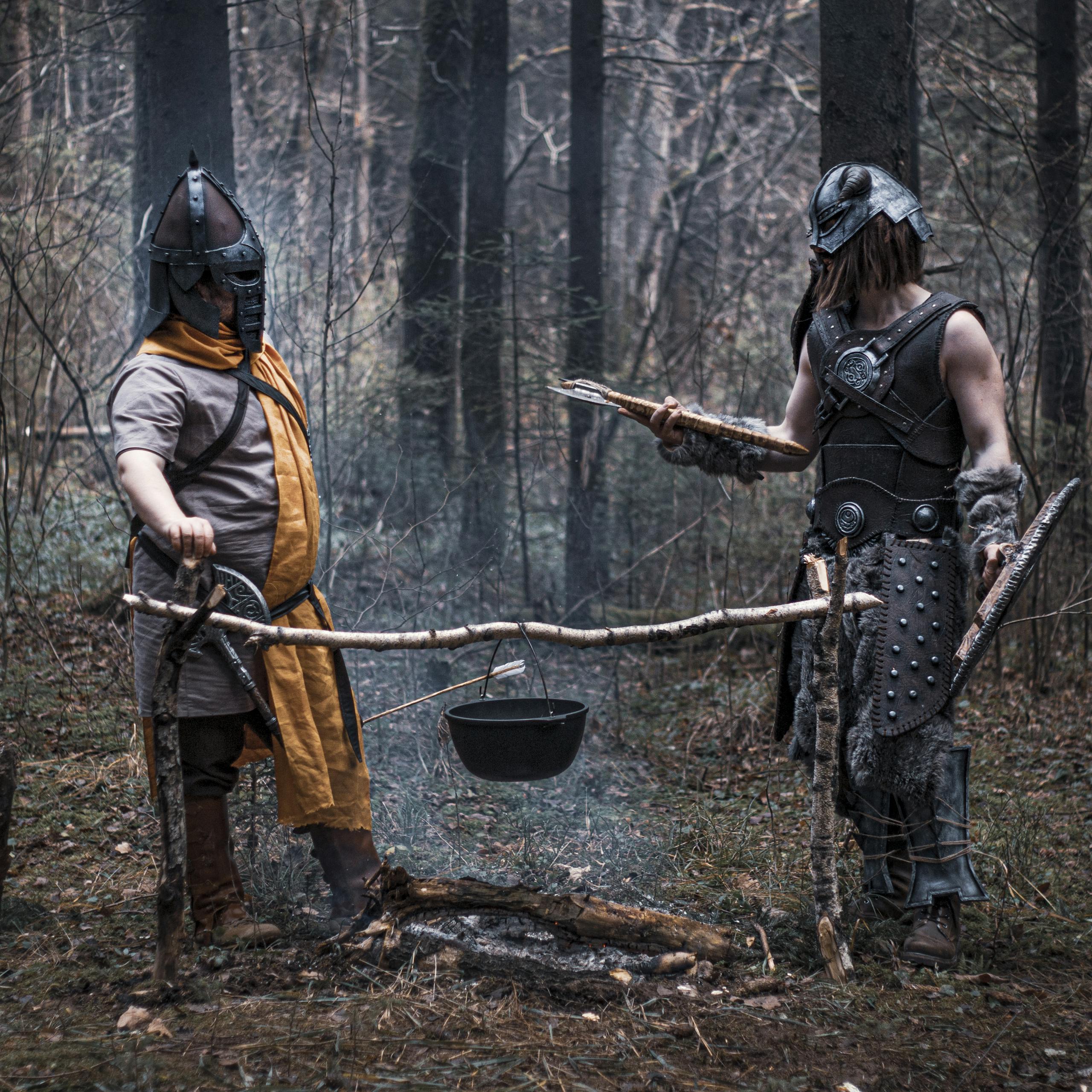 Two medieval reenactors cooking over a bonfire in a dense forest, dressed in historical armor.