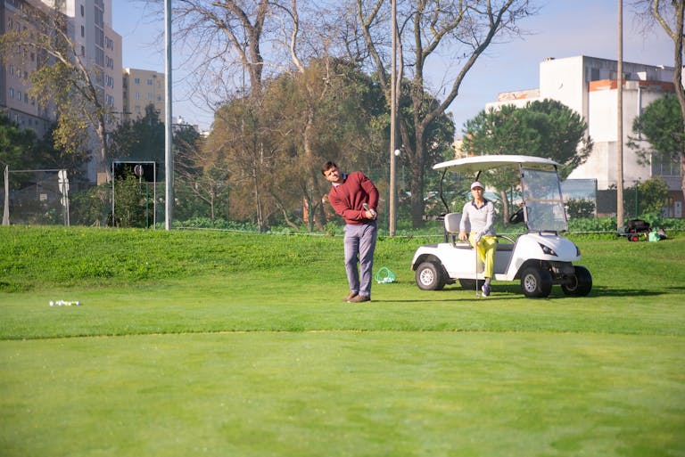 Two adults playing golf on a sunny day, using a golf cart at a lush green course.